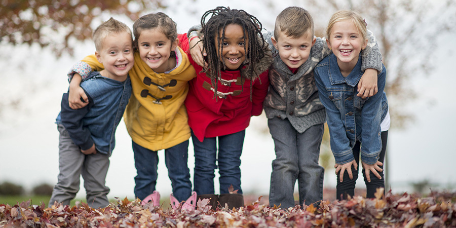 A multi-ethnic group of elementary age children are playing outside on a fall day. They are standing in a row and are smiling while looking at the camera.