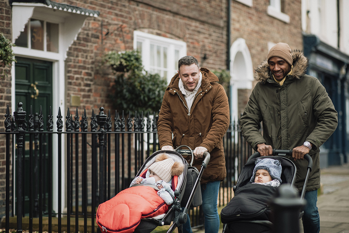 Two male friends out walking on a sidewalk and pushing their babies in strollers.