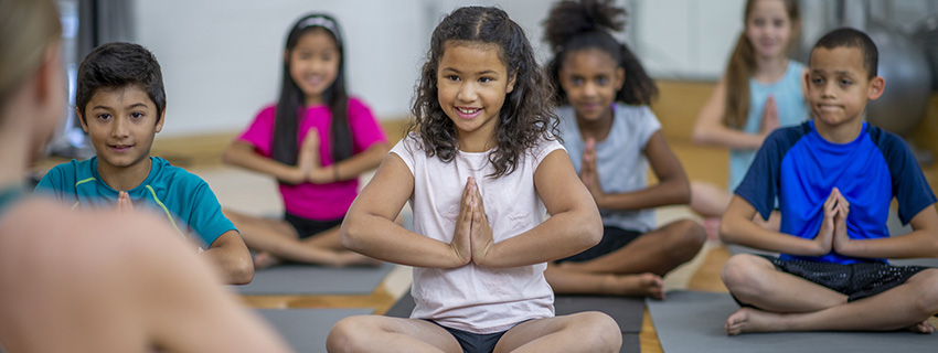 A teacher is leading her students during physical education class. She is meditating with her hands together, and her students are following her