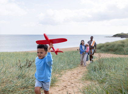 A wide shot of a joyful family walking up a sandy coastal path, surrounded by sand dunes at a beach