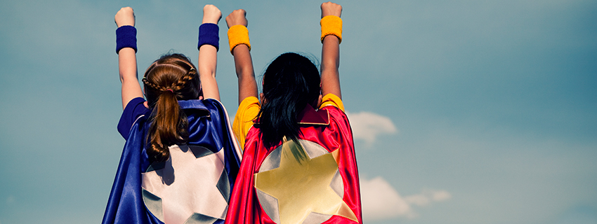 Two girls in superhero capes hold their arms above their heads. 
