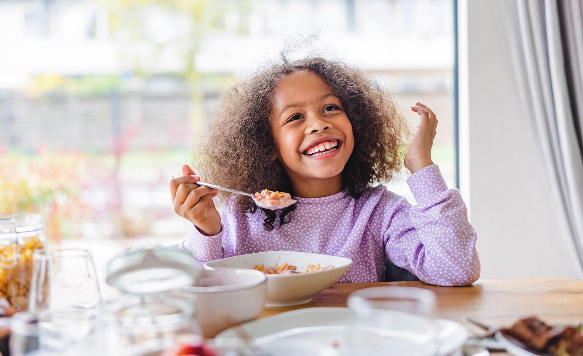 Young girl eating cereal with a smile at the table.