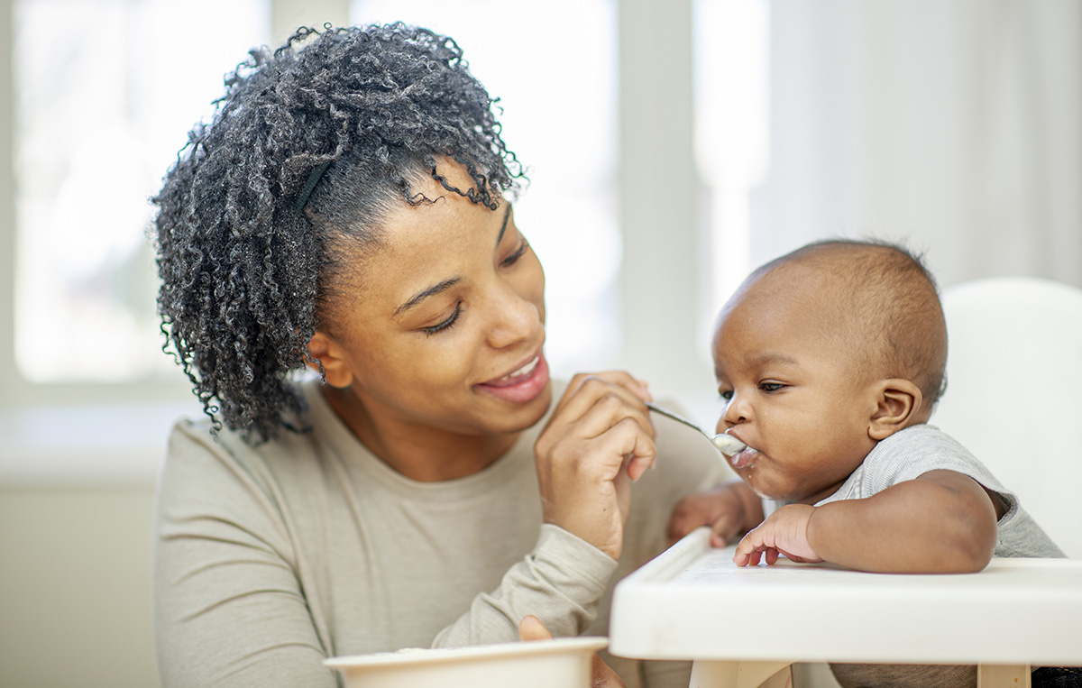 A young mother of African decent sits beside her infant son as he sits in his highchair and she feeds him.