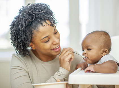 A young mother of African decent sits beside her infant son as he sits in his highchair and she feeds him.