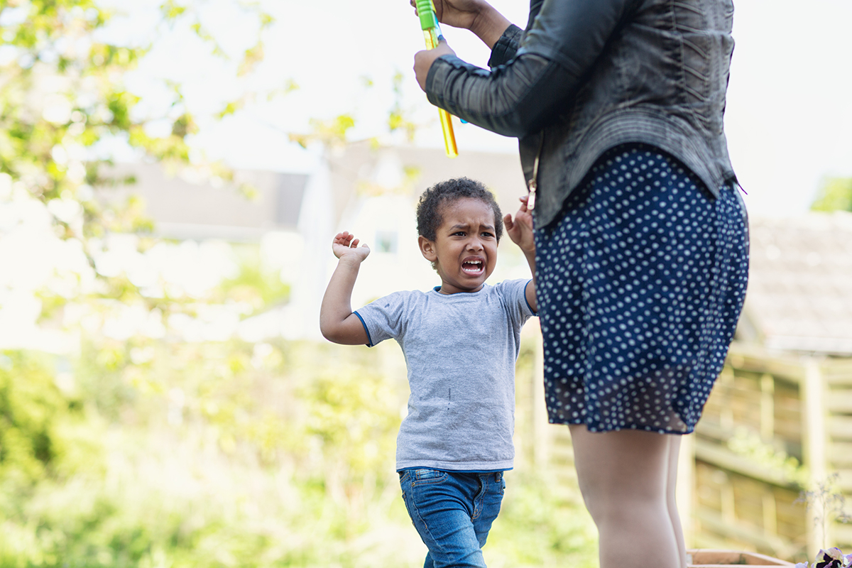 A toddler boy screaming ad crying in disappointment about something his mother will not do. Mother is there too, but focus is on the boy. They are outdoors in backyard, it is summer